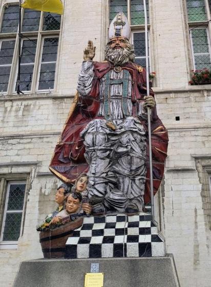 Auf dem Bild sieht man die bunte große Statue von #Saint Niklass vor dem #Rathaus sitzen. Zu seinen Füßen sind links drei Kinderfiguren und ein Sack zu sehen.  Nikolaus  segnet mit seiner rechten Hand und hält in seiner Linken den Bischofsstab.