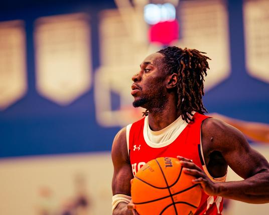 A black man with dreadlocks looks to the left while holding a basketball in front of himself,  he's wearing a red tank top and behind him you can see a bit of the basket backboard and a countdown timer as well as an array of white chevrons on a wide blue stripe on the wall