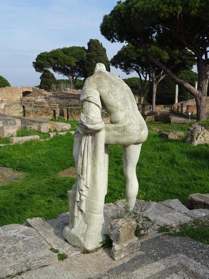 Auf Mauerresten steht eine Steinstatue - eine nackte Person von hinten mit einem Bein hoch auf ein Fragment gestützt, der Kopf fehlt. Dahinter Grasfläche und dahinter Ruinen und Steine mit Pinienbäumen.