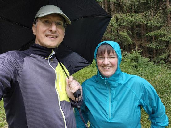 In this image, we see a man and a woman standing together in the woods. The man is wearing a sun hat and glasses, while the woman is wearing a blue jacket and holding an umbrella. They are surrounded by trees, grass, and plants. The dominant colors in the image are black and a vibrant blue accent color. Both individuals are smiling, with the man standing slightly behind the woman. The woman's face is partially obscured by the umbrella she is holding. The scene conveys a sense of outdoor leisure…