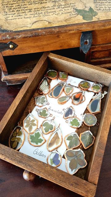 English A small wooden cabinet with one drawer open. Inside are glass jewels containing pressed leaves and insect wings. Only part of the silver-finish soldering remains to be done. It is noted that there are earrings, bracelets and necklaces.
Français Un petit meuble en bois dont un tiroir est ouvert. Dedans il y a des bijoux en verre contenant des feuilles pressées et des ailes d'insectes. Il ne reste qu'une partie de la soudure finition argentée à terminer. Il est noté qu'il y a des boucles d'oreille, des bracelets et des colliers.