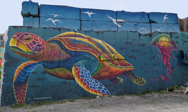 The image features a vibrant mural painted on a concrete wall. The mural depicts a large, colorful sea turtle swimming to the left, with its shell displaying a spectrum of colors including red, yellow, green, and blue. The turtle's flippers are also multicolored, with shades of blue, red, and yellow. To the right of the turtle, there is a jellyfish with a pink and yellow bell and long, flowing tentacles. The background of the mural is a deep blue, representing the ocean, with silhouettes of fis…