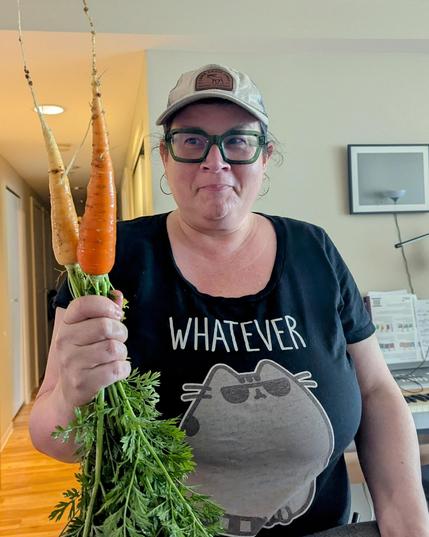 A woman standing inside an apartment side-eyes two large carrots (with greens still attached) that she's holding. The woman is wearing a black Pusheen t-shirt that says "Whatever". 