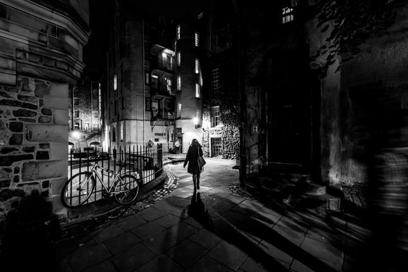 Schwarzweissfotografie einer nächtliche Szene in einer engen, gepflasterten Gasse mit alten Gebäuden. Eine Person mit Mantel und Tasche läuft von der Kamera weg in eine schwach beleuchteten Hinterhof, das Licht wirft ihren langen Schatten auf den Boden. Links hängt ein Fahrrad an einem Eisenzaun. In den Fenstern der Gebäude brennt Licht.
