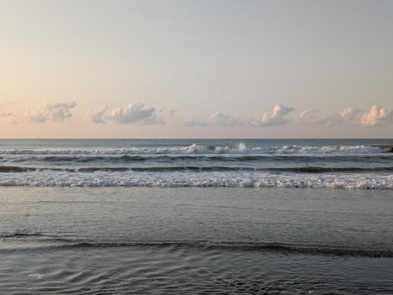 Photo looking out over the ocean. There is a line of shallow water with ripples followed by a line of breaking waves and finally the distant ocean all the way to the horizon. What appears as a single line of puffy white clouds tinged with pink is just above the horizon. The sky is clear and light blue otherwise, with a touch of pink toward the left of the photo, where the sun just rose.