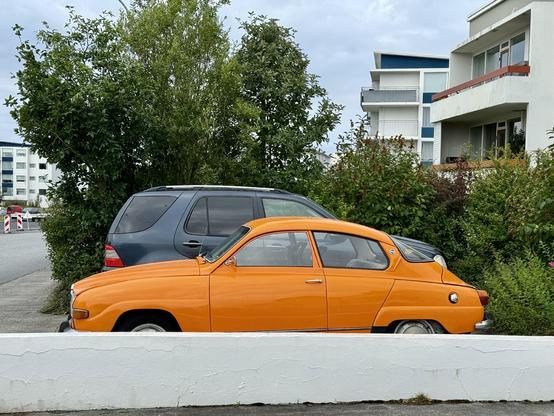 An orange Saab 96 parked next to a low wall