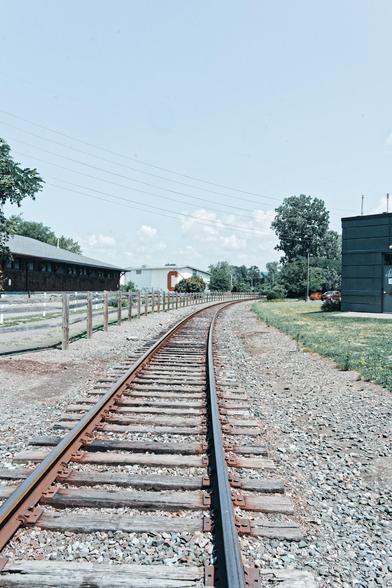 Railroad tracks receed into the background and curve to the right as they go out of sight.  On the left side is a big brown building with a split rail fence in front and behind that a low white building with a red C on it,  on the right a grassy lawn and a little bit of a dark colored building,  some powerlines cross the skyu which is blue except for some cumulus clouds in the distant background