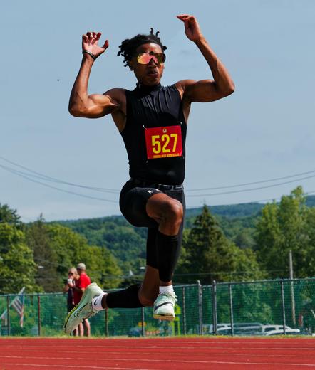 Black man with dreadlocks wearing reflective glasses hangs in the air with a red track, fence, and spectators in the background and past that the roofs of some cars,  a close row of trees and a distant forest as well as power lines.  He's got his arms up in the air,  a black tank top that shows his muscular arms, one leg bent in front of him and one behind, nike socks and shoes and a red bib that says 527 in yellow numerals.