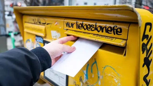 Eine Hand steckt einen Brief in einen gelben Briefkasten mit dem Graffiti „Nur Liebesbriefe“ darüber.