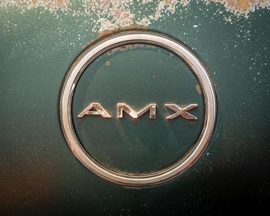Color photo of the AMX badge on a green AMC Javelin AMX. The badge and surrounding body paint are pitted and show signs of rust. 