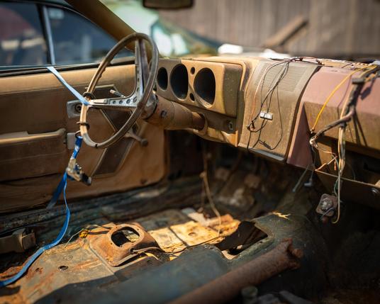 Interior of an AMC Javelin AMX. Pretty much gutted with no seats, instruments, etc. The driver’s side door is held closed with a bungee cord looped around the steering wheel.