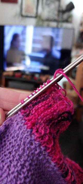 In the foreground is some knitting being held in my left hand close up to the camera. The knitting is on two fine metal needles, and is purple on the left and a stripe cerise and black on the right. In the background is a large, out of focus TV showing an episode of Star Trek DS9.