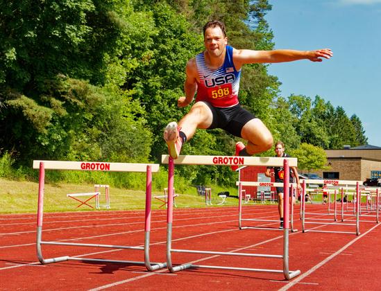 A man jumps over the rightmost (his left) hurdle of a pair,  both of which have a white bar with the word GROTON on them.  He's got a red, white and blue shirty with irregular stripes and has one leg in front of him and the other arm out straight to your right with other leg and arm folded back and another runner visible in the far background