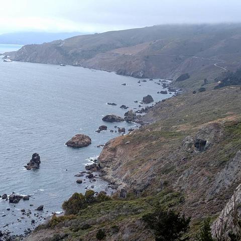 A rugged stretch of California coastline is seen from above, with steep, rocky cliffs descending into the Pacific Ocean. Large boulders and sea stacks dot the shoreline and water, while the coastal hills in the distance are partially shrouded in mist. The muted tones of the overcast sky lend a soft, atmospheric quality to the scene. Sparse vegetation covers the cliffs, adding texture to the dramatic landscape.