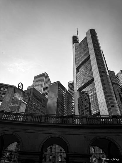 Schwarzweißaufnahme aus Frankfurt am Main. Im unteren Bildbereich alte, steinerne Rundbögen, darüber ragt eine moderne Skyline mit Hochhäusern empor. Die Perspektive ist steil nach oben gerichtet, was den Kontrast zwischen historischer Architektur und moderner Baukunst betont. Der Himmel ist leicht bewölkt, das Licht weich und zurückhaltend