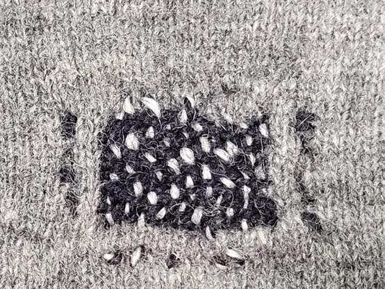 Close up of a light grey knit sweater showing a multi-thread, woven repair patch