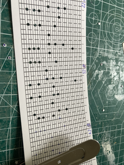 Auf einer Arbeitsunterlage liegt ein Lochband für eine 30 Ton Spieluhr. Darauf ein Lochstanzer. In einem Teil des Bandes befinden sich bereits Löcher.