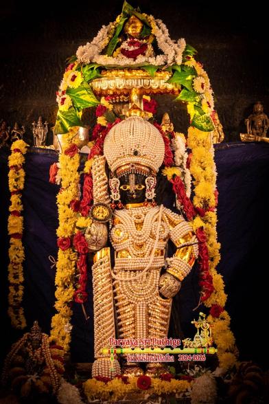 Idol of Hindu deity Krishna with a "chain mail" of pearls with gold undergird, standing with a matching dome-shaped crown and a similarly clad staff in right hand. A string of red roses surround Krishna. Loose yellow and red flowers at Krishna's feet. A gold umbrella over Krishna. A string of yellow beetle leaves over the umbrella from left to right. Strings of white flowers atop the umbrella. Long strings of yellow flowers hang down from the umbrella: one on either side of Krishna. Dark cloth background.