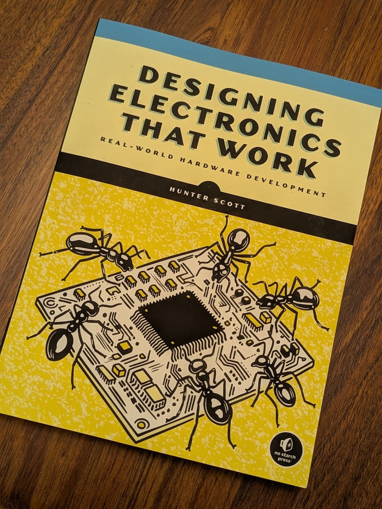 a physical copy of the book, "Designing Electronics That Work." "Real-World Hardware Development" by Hunter Scott there's an illustration of a small square circuit board being attended by six ants.