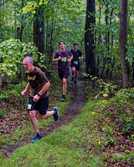 Three runners in the rain on a trail that starts out in the far middle ,  goes between two trees with rectangular blazes,  then curves to exist on the left.
