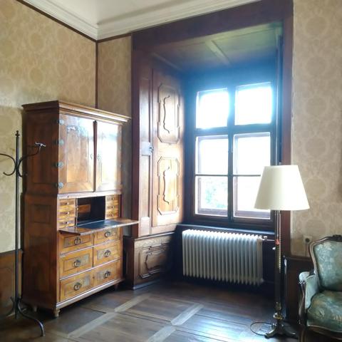Mächtiges Fenster in einer dicken Wand. Daneben steht ein Sekretär mit vielen Schubladen, der mit Intarsien verziert ist.