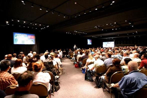 come together at events: a photograph of a traditional conference. A sea of attendees sit facing toward a distant standing speaker.