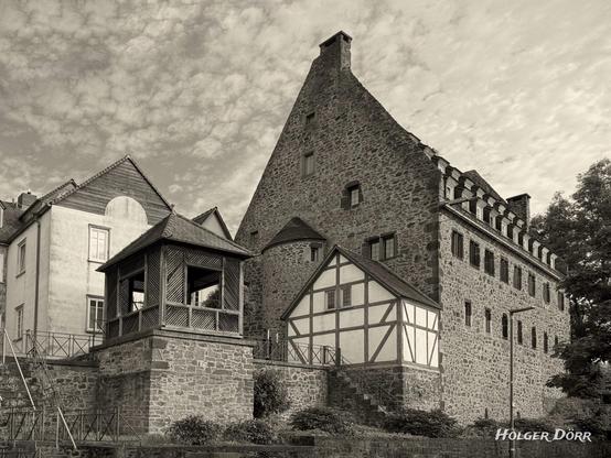Sepiaaufnahme des Barfüßerklosters in Grünberg: massiver Steinbau, Pavillon und Treppe im Vordergrund, daneben Fachwerk; darüber strukturierte Wolken - ruhige, zeitlose Stimmung.