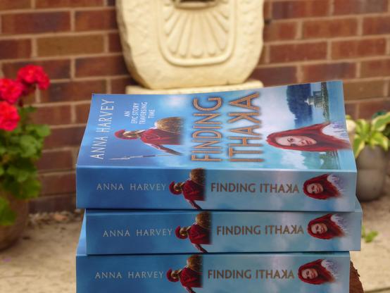 Copies of Finding Ithaka, with distinctive blue cover, in front of a fountain,  potted plant and geranium. 