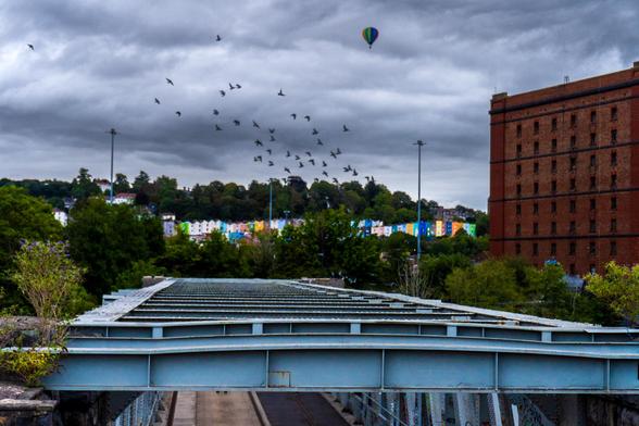 Looking over the metal slats over a bridge with colourful terraced houses in the background, a swarm of birds diving diagonally from top left to bottom right in the middle of the pic and a single hot air balloon at the top of the pic slightly right. The right side is a big red brick building. The sky is quite grey and cloudy.