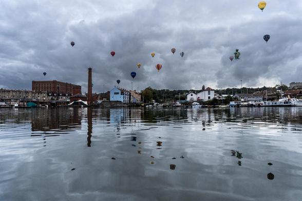 Looking over the water to a Dock area with a red brick building on the left with smokestack ans some houses and boats docked all along. The sky is grey and cloudy and there are 10-15 hot air balloons in the sky. You can see distorted reflections of the balloons in the water which is also grayish blue.