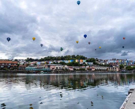A different view over the water with a blue bridge and colourful terraced houses on a hill across the water. Again cloudy sky and 10-15 hot air balloons scattered through the sky.