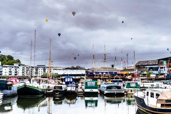 Boats parked in a marina with a cloudy sky and many hot air balloons, mostly quite distant. Just the very top of Bristol cathedral is also visible.