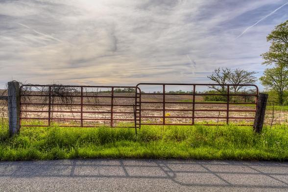 A rusty metal gate stands between lush green grass and a plowed field, under a vast, cloudy sky.