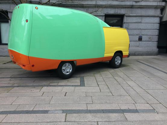 The image features a uniquely designed van parked on a paved area in front of a stone building. The van has a large, rounded body with a mint green color on the upper half and a bright yellow on the lower half, separated by an orange band near the bottom. The rear of the van is mostly mint green, with a small section of yellow at the back. The van has two visible wheels, with silver rims, and red tail lights. The building behind the van has a classical architectural style with large windows and…