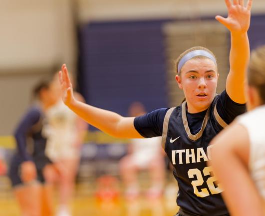 A woman wearing a blue jersey that reads ITHACA 22 and a light blue headband holds one arm up and the other to the side to block another women who can be seen blurred out on the far edge of the frame while other players are blurred out in the background