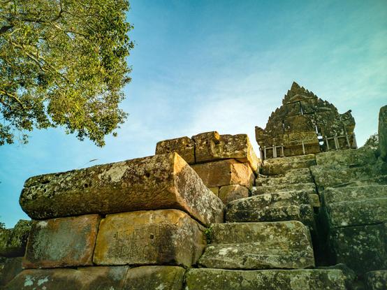 Photo shows the stoney stair steps upwards to the entrance gate to a temple. The entrance gate has a triangle top form, with small triangle tips on its side.  The colours are light brown and red stone, and the large stepping stones in the stair is grey. There are some intricate decorations on some of the stones. The photo is taken angled upwards, having the mostly blue sky as the main background element. To the left there is the top of a large, green tree framing the steps and the temple entrance.