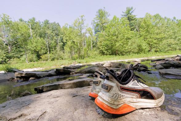 In the foreground a pair of Puma sneakers mostly white with black heels and a black insert on the side outliner in red;  these are on a rock and in the background find a wide creek with lots of rocks and a green forest on the other side