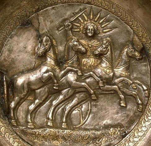 A happy, smiling Helios is driving his horses with a riding crop on the relief on a silver kylix. The horses are prancing over a flowery medow, drawing he sun wagon behind them. Helios is wearing a gilded chiton and cloak and a nine-spiked sun ray crown.