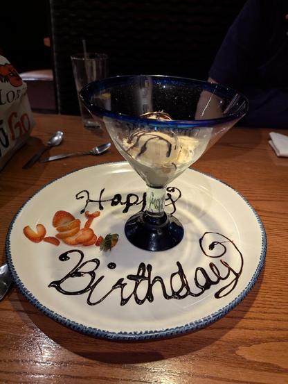 Scoop of vanilla ice cream with a chocolate drizzle, sitting in a far too large cocktail glass atop a plate. The plate says "Happy Birthday" in chocolate drizzle and is adorned with a sliced strawberry arranged in the shape of a lobster.
