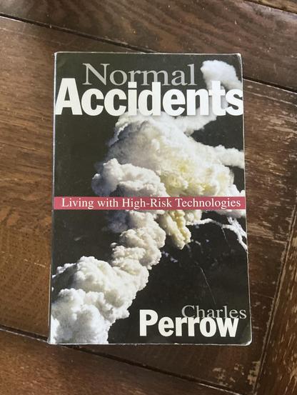 A black paperback book on a wood table with an image of the cloud of the Challenger explosion, at the top the title Normak Accidents, a red bar white white letters across the middle says Living with High-Risk Technologies, and the author’s name Charles Perrow
