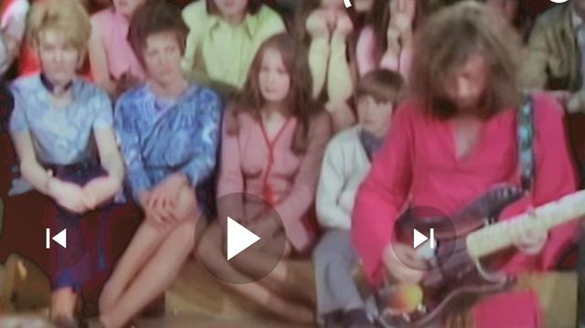 Szene aus einer TV Sendung, aus 1970, mit einem Mann, der eine Gitarre spielt. Er trägt ein leuchtend rotes Gewand und hat langes, lockiges Haar. Die Gitarre hat einen dunklen Körper mit einem gelben Streifen. Im Hintergrund sitzen mehrere Personen, die in verschiedenen Farben gekleidet sind, darunter Blau, Pink und Grün. Die Personen im Hintergrund scheinen aufmerksam zuzuschauen. 