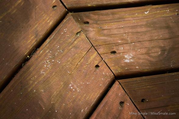 Brown wooden planks are arranged in a geometric pattern, with visible screw holes and minor wear. Small white specks are scattered across the surface.