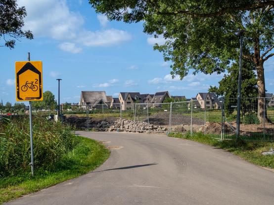 Next to a winding path is a yellow sign depicting a bicycle with a superscript number two. In the background, the new residential buildings of Weespersluis, Weesp, The Netherlands. Lush greenery is visible on either side of the path under a blue sky with a few scattered clouds