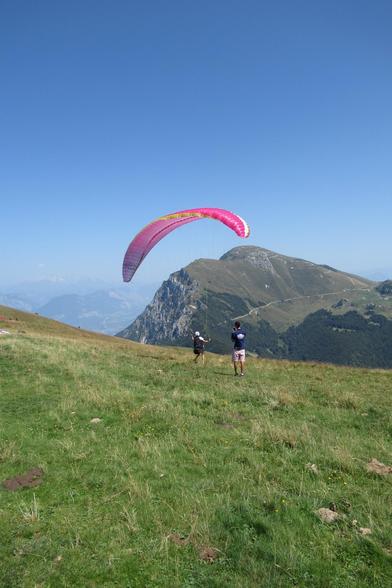 A person is paragliding with a red paraglider, going down a hill, with another person next to them. In the foreground is a vast green field, with mountains at the back