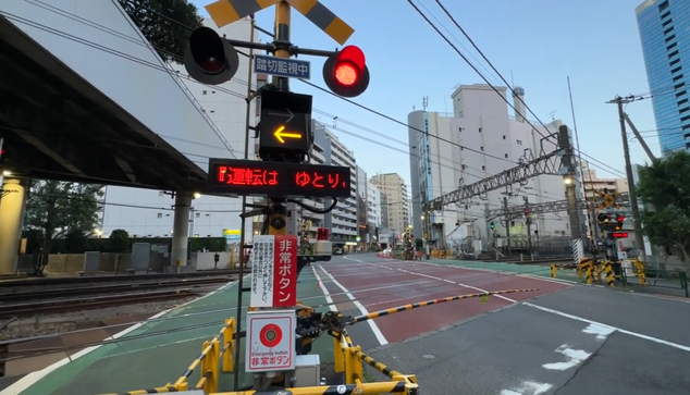 Japonia, Shinjuku. Przejazd kolejowy. Opuszczone rogatki. Po obu stronach drogi zapalone jedno czerwone światło z dwóch migających naprzemiennie. Wyświetlacze wskazują kierunek, w którym przejedzie pociąg (z prawej do lewej), pod nimi przyciski bezpieczeństwa awaryjnego zatrzymania pociągów z tablicą ostrzegawczą, syreną i lampą.