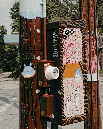 Decorated electric box and pole.  The pole has an oval portrait of the character Totoro and is painted brown like a tree except for being sky blue near the top.  The electric meter is whte but it's attached to one of two small brown boxes behind a big brown box which has some text written on it and has 3 of those little black things and lots of sakura petals on top and bottom and yellow stripe in the middle and two other abstract characters.