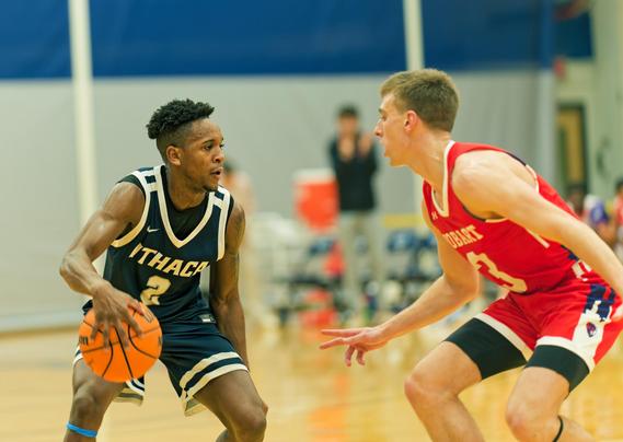 A black man with natty hair is wearing a blue jersey with the number 2 and has the basketball and looks to the right where a white man two inches taller confronts him wearing a red jersey that says 3,  in the background you can see a two-toned blue and white gynanisum ball and a orange Gatorade-branded container with a blurred out man who seems to be in charge of it.