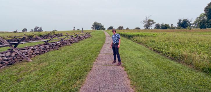 A wide picture with an almost straight path that curves to the left in the distance with a young man wearing dark grey pants and a blue and white shirt standing on one side of it.  To the right there is a strip of close mowed grass and then a field of taller grass with a few psoradic trees,  to the right close cut grass with stone walls and primitive rail fences