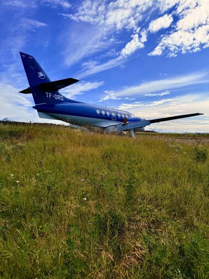 A British Aerospace Jetstream 32 with registration number TF-ORD sits disused at RKV airport. The sky is blue with whisky white clouds and ground is grass covered. The plane has Eagle Air Iceland livery. 