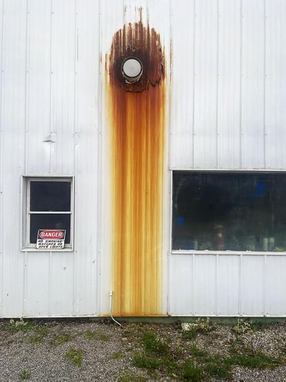 A weathered metal wall displays prominent rust stains beneath a vent, flanked by two windows, one of which has a danger sign
